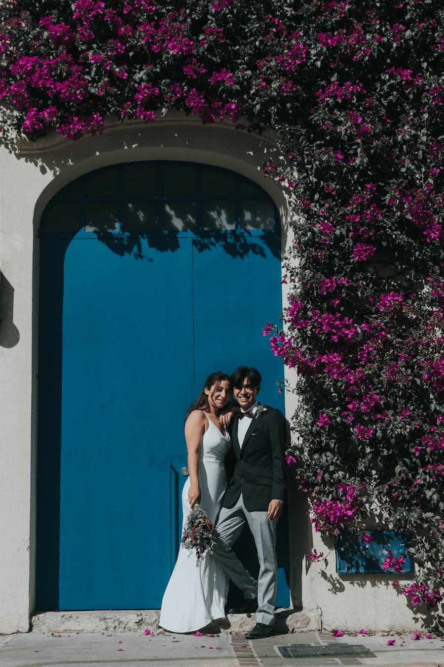 Novios posando el día de su boda frente a una puerta de madera en una pared de bugambilia.